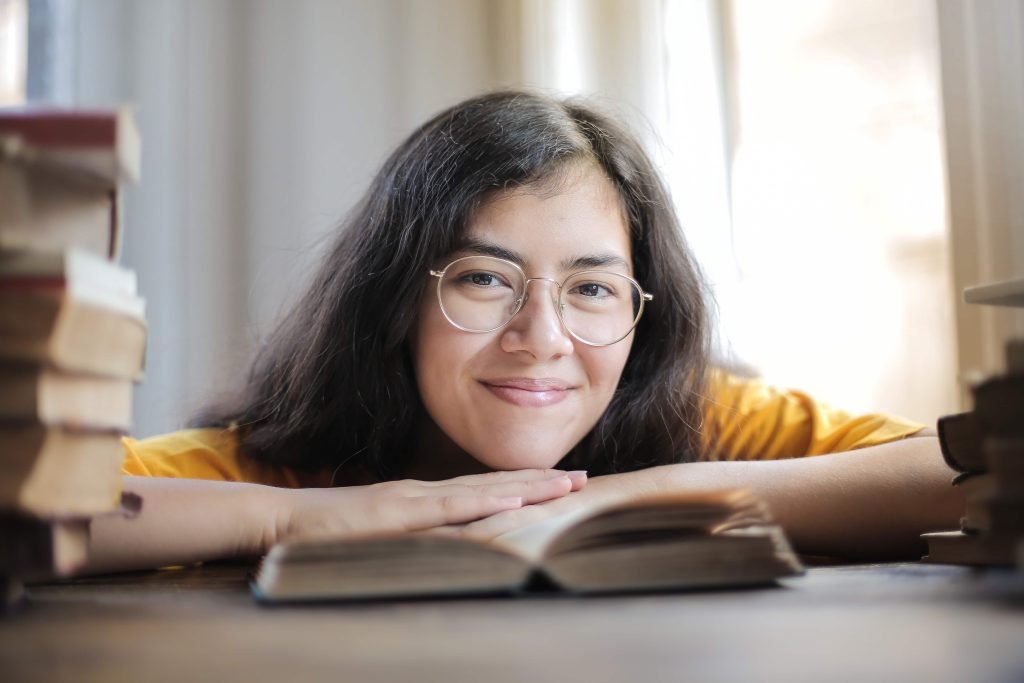 Jeune femme asiatique à lunettes qui sourit, menton sur les mains, au milieu de livres